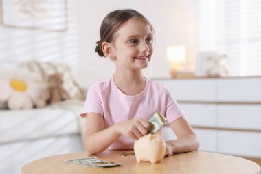 Küçük kız, ahşap masadaki domuz kumbarasına cep harçlığı koyuyor.