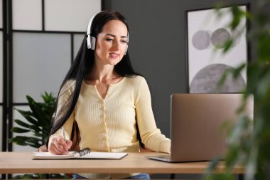 Kulaklıklı bir kadın, bilgisayarla kapalı bir masada çevrimiçi sınavdan geçerken not alıyor.