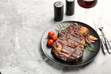 Delicious grilled beef steak with rosemary, onion, tomatoes, cutlery and shakers on grey textured table, closeup. Space for text