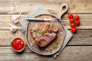 Delicious grilled beef steak with rosemary, spices, ketchup, carving fork and products on wooden table, flat lay