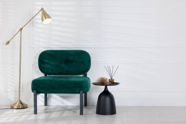 Stylish lamp, comfortable armchair and side table with decor near white wall indoors