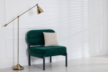 Stylish lamp and comfortable armchair with cushion near white wall indoors