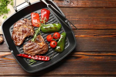 Pan with grilled beef steaks, rosemary, vegetables and carving fork on wooden table, flat lay. Space for text