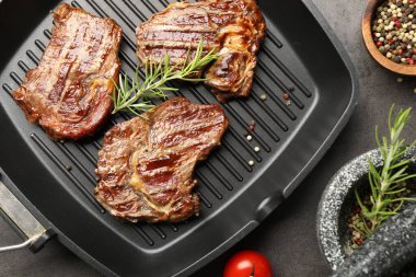 Pan with grilled beef steaks, rosemary and spices on grey textured table, flat lay