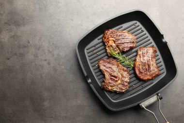 Pan with grilled beef steaks, rosemary and peppercorns on grey textured table, top view. Space for text