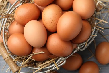 Raw chicken eggs, metal basket and straw on grey wooden table, flat lay