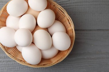 Raw chicken eggs in wicker basket on grey wooden table, top view. Space for text