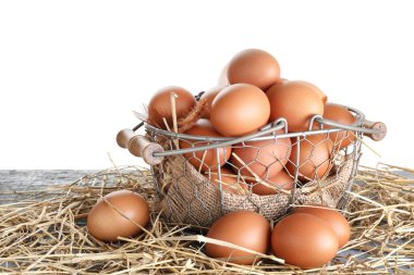 Raw chicken eggs, metal basket and straw on grey table against white background