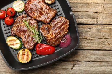 Pan with grilled beef steaks, rosemary and vegetables on wooden table, top view. Space for text
