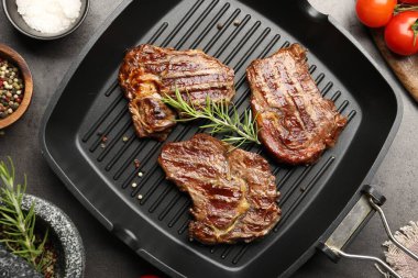 Pan with grilled beef steaks, rosemary and spices on grey textured table, flat lay