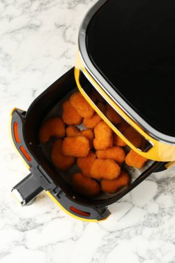 Air fryer container with cooked chicken nuggets on white marble table, top view
