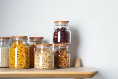 Beyaz duvarın yanındaki ahşap masada farklı ürünler bulunan cam kavanozlar. Metin için boşluk