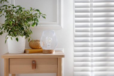 Şık bir lamba, hasır depolama kutusu, komodinin üstünde beyaz duvarın yanında kitap ve çiçek. Metin için boşluk