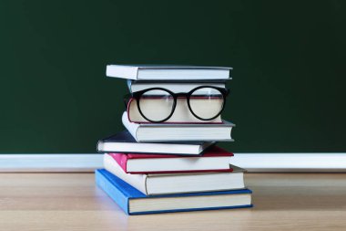 Öğretmenler Günün kutlu olsun. Tahtanın yanındaki tahta masada kitap ve gözlük yığını.