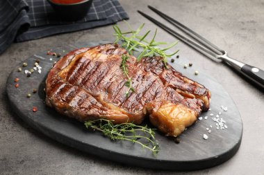 Lezzetli ızgara biftek, otlar, baharatlar ve çatal, gri desenli masa, yakın plan.