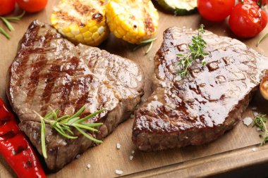 Delicious grilled beef steaks with herbs and vegetables on wooden board, closeup