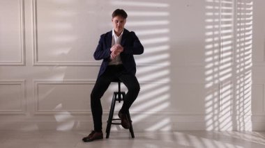 Şık kotlu bir adam beyaz duvarın yanındaki taburede saati kontrol ediyor.