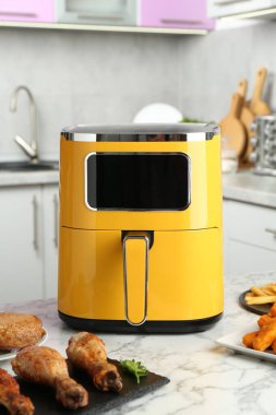 Modern air fryer and cooked meals on white marble table in kitchen