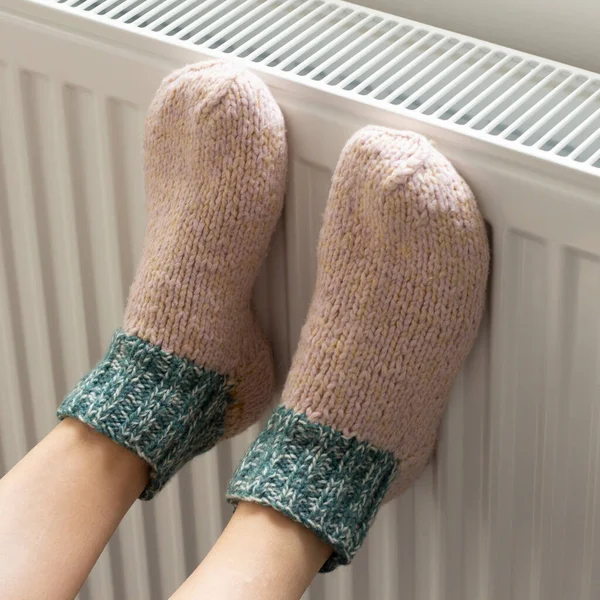 A woman warms her feet in woolen socks on a radiator. Home heating. Low temperature and cold in the house. Photo closeup