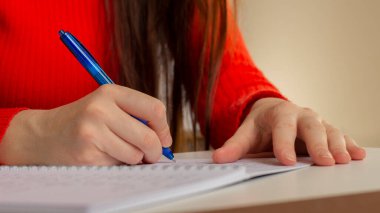A girl at the table writes with a pen in a notebook. Student studying, taking notes. School and education. Closeup photo