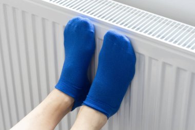 A woman warms her feet in woolen socks on a radiator. Home heating. Low temperature and cold in the house. Photo closeup