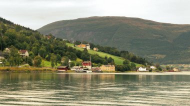 Olden, Norveç 'te Innvikfjorden, 15 Ağustos 2024 - Yuvarlanan tepeler ve sakin fiyortlar arasında pitoresk bir kıyı köyü. Renkli evler manzarayı noktalıyor