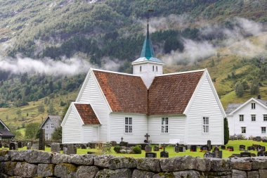 Eski Norveç Kilisesi, Norveç 15 Ağustos 2024 - Çan kulesi olan tuhaf beyaz bir kilise, yuvarlanan tepeler ve sisli dağlarla çevrili huzurlu bir mezarlığın ortasında duruyor