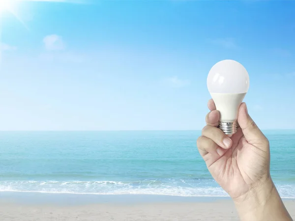 Hand holding a LED light bulb on a sandy beach with clear sky. Using economical and environmentally friendly light bulb concept