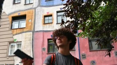 Viyana, Avusturya, Ağustos 2022. Beyaz bir gencin, arkasında Viyana 'daki Hundertwasser evinin renkli yüzüyle selfie çekerken güzel bir yavaş çekim görüntüsü. Konut, kondominyum.