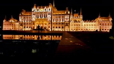 Budapeşte, Macaristan, Ağustos 2022. Parlamentonun ikonik görüntüsüyle baş döndürücü gece görüntüleri, geniş açılı mercekle alçak açı görüntüsü. İnsanların siluetlerini tanırsın. Çeşmedeki yansıma.