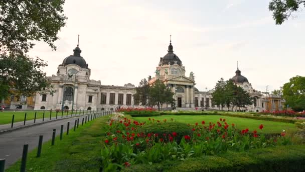 ハンガリー ブダペスト 2022年8月 最大のSzchenyi風呂への入り口で素敵なパンの映像 花のある庭園は美しい建築物のフレームです 人々が出入りしている — ストック動画