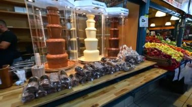 Budapest,Hungary,August 2022. At the central market pan footage on the counter of Kurtoskalacs, the typical local sweet. Next to the desserts, two cascades of liquid chocolate, one white and one black