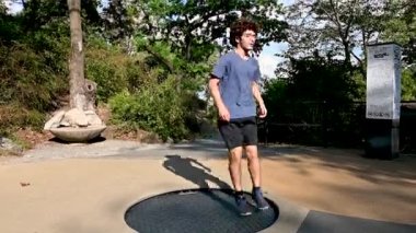 A young Caucasian man with curly hair and glasses is having fun jumping on a trampoline in a public park. Youth and freedom carefree concept.