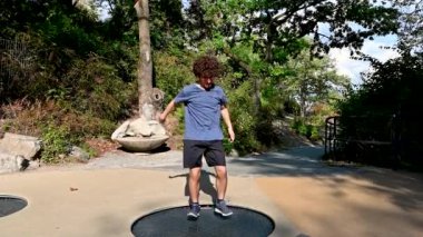 Slow motion footage of a young Caucasian man with curly hair and glasses jumping having fun on a trampoline in a public park. Youth and freedom carefree concept.