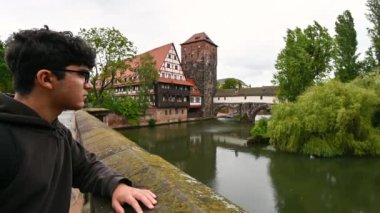 Nuremberg, Almanya, 1 Ağustos 2023. Genç beyaz bir adam, nehrin büyüleyici manzarasına hayran kalıyor. Yarı ahşap ev suya yansıyor. Nuremberg 'i keşfetmek ve keşfetmek.
