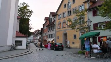 Wangen im Allgu, Almanya, 17 Ağustos 2023. Mavi saatte çekim, eğim hareketi çan kulesini vurguluyor, bir araba geçiyor. Aydınlatılmış dükkan pencereleri.