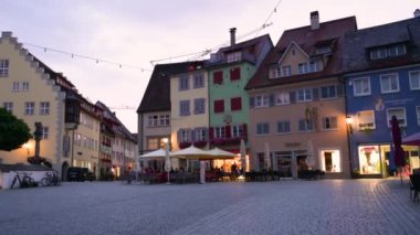 Wangen im Allgu, Almanya, 17 Ağustos 2023. Mavi saatte çekim, eğim hareketi çan kulesini vurguluyor, bir araba geçiyor. Aydınlatılmış dükkan pencereleri.