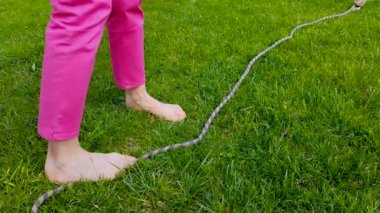 Beyaz bir kadının ayakları üzerinde çıplak ayakla yürüdüğü bir yakın çekim görüntüsü: sadece ayaklarıyla çimlerin üzerinde dinlenen bir halatı yakalamaya çalışır. Güzel güneşli bir gün.