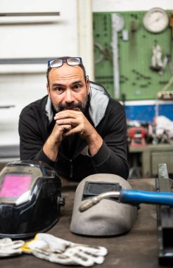 Kameraya bakan beyaz bir adamın güzel bir portresi. Yedek kulübesinde kaynak aletleri var: iki koruyucu kask ve kaynak tabancası. Dikey biçim.