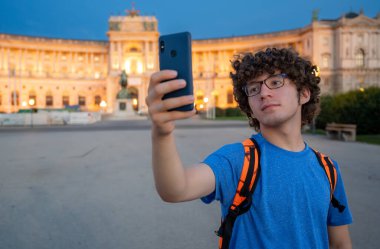 Vienna, Austria, August 17, 2022. A young Caucasian man with curly hair and glasses takes a selfie in front of the Hofburg Imperial Palace at sunset. The warm evening light enhances the imposing facade. clipart