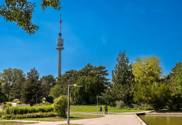 stock image Vienna, Austria, August 17, 2022. The Danube Tower or Donauturm is the tallest building in Austria at 252 metres. It stands out among the trees of the park, three tourists walk along the avenue.