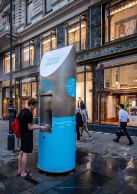 Viyana, Avusturya, 18 Ağustos 2022. Eski kasabadaki bir sokakta içme suyu dağıtıcısı şişesini suyla dolduruyor. Yan taraftaki işaretler içecek servisinin su püskürttüğünü gösteriyor.