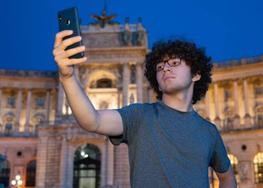 Viyana, Avusturya, 18 Ağustos 2022. Kıvırcık saçlı ve gözlüklü genç bir beyaz adam mavi saatte Hofburg İmparatorluk Sarayı 'nın önünde selfie çekiyor. Işıklandırma etkileyici dış görünüşü geliştirir..