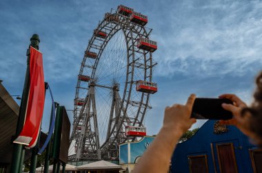 Vienna, Austria, August 18, 2022. Shot of the large vintage ferris wheel in the city's amusement park, the prater. A mobile phone is lifted to take a photo. Use of technology. clipart