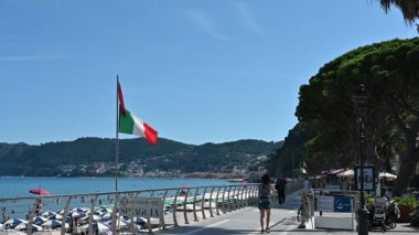 Alassio, İtalya, 14 Temmuz 2024. Deniz kenarındaki gezinti alanının ikonik yavaş çekim görüntüleri. İtalyan bayrağı rüzgarda dalgalanıyor, insanlar kaldırımda yürüyor. İtalyan deniz kıyısında rahatlama konsepti.