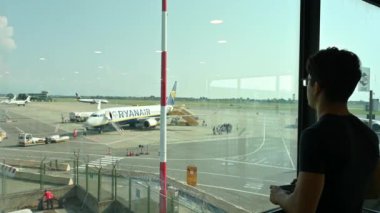 Caselle Havaalanı, Torino, İtalya. 13 Ağustos 2024. Kafkasyalı genç bir adam pistte pencereden dışarı bakar ve cep telefonuyla bir Ryanair uçağının fotoğrafını çeker. Seyahat yaşam tarzı ve teknoloji kullanımı.