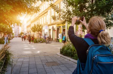 Viyana, Avusturya, 23 Ağustos 2022. Sırt çantalı orta yaşlı beyaz bir kadın eski kasabadaki bir caddede cep telefonuyla fotoğraf çekiyor. Teknolojiyi kullanarak seyahat et..
