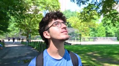 New York, ABD, 14 Ağustos 2024. Central Park 'ta yürüyen kıvırcık saçlı ve gözlüklü genç beyaz bir adamın portresi etrafa bakıyor ve gördüklerine yorum yapıyor. Sağlıklı açık hava yaşam tarzı.