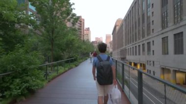 New York, ABD, 14 Ağustos 2024. POV görüntüleri yüksek hat boyunca yürüyor: genç beyaz bir adam önümüzde, yeşil ağaçların arasında yürüyor. Aşağıdan geçen arabaları görebiliriz..
