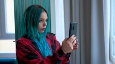 Pencerenin önünde durup cep telefonuyla selfie çeken güzel mavi saçlı beyaz kadın portresi. Boş zaman ve teknoloji kullanımı, sosyal içerik oluşturma.